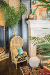 An interior with a potted palm, a wicker chair, blue walls, a traditional fireplace mantel, and modern artwork in colors that include the new 2024 Pantone Color of the Year, Peach Fuzz