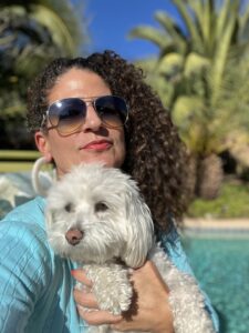 Color strategist Lauren Battistini wears a turquoise top and holds a little white dog in front of palm trees