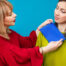 A blonde woman in a red shirt holds a blue color sample up to the face of a brunette woman in a chartreuse blouse in order to test her color compatability. Both are against a turquoise background
